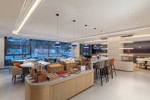 A modern cafe with a central counter offering various food items, surrounded by seating areas with large windows providing views of the outside.