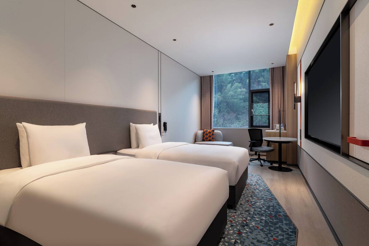 A hotel room with two beds, a desk, a chair, and a large window.