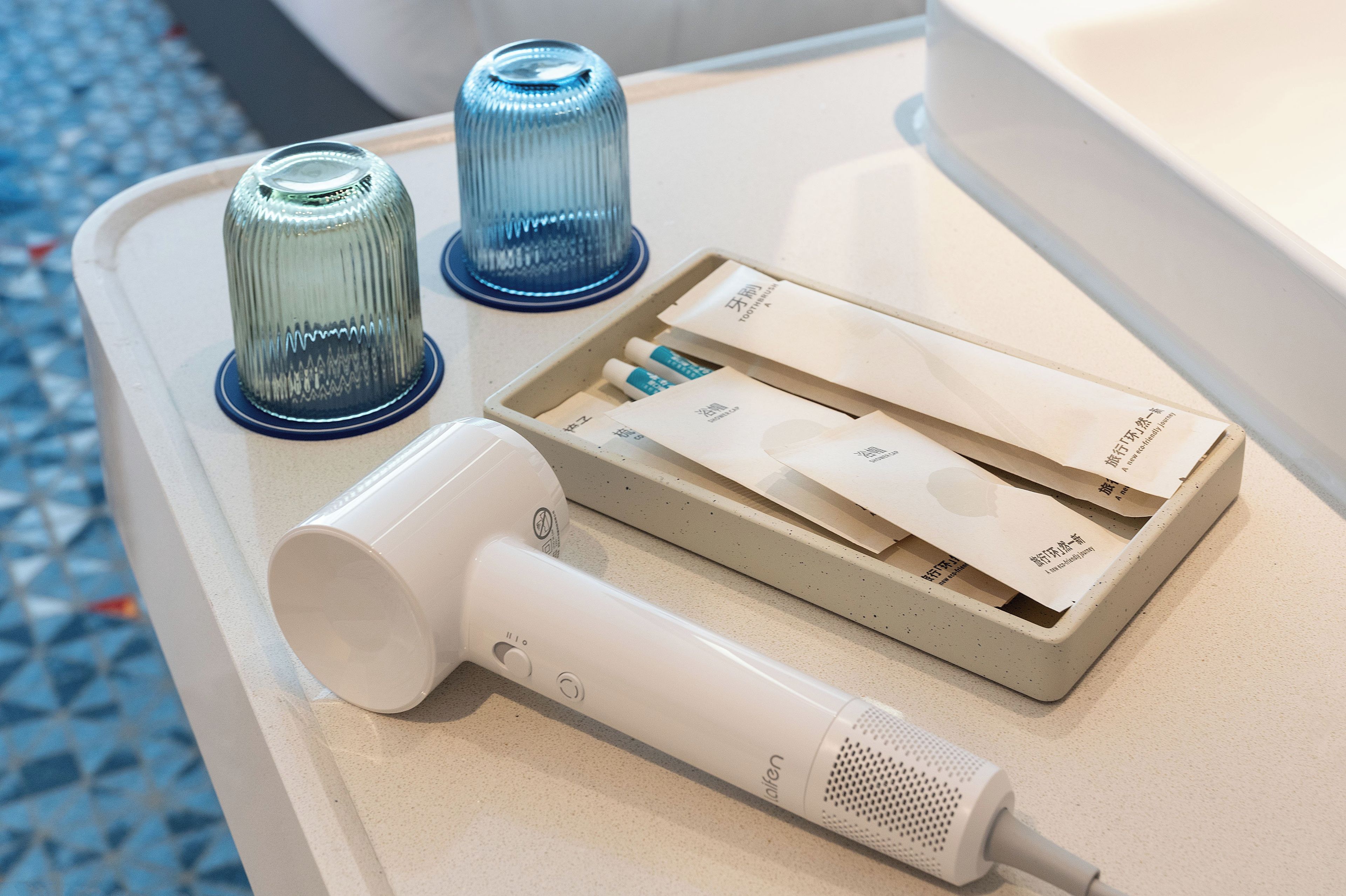 A hotel room with a white bedside table, a white hairdryer, a blue glass tumbler, and a tray with a tissue box and a small item.