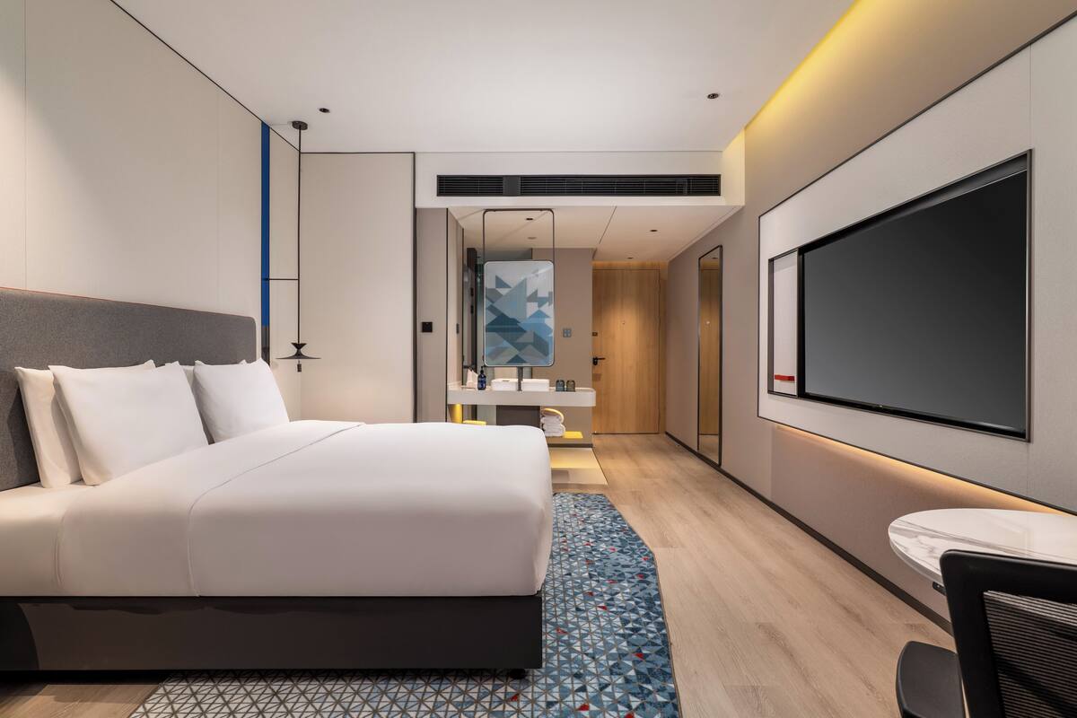 A modern hotel room with a large bed, a TV, a small table, and a bathroom with a mirror and sink.