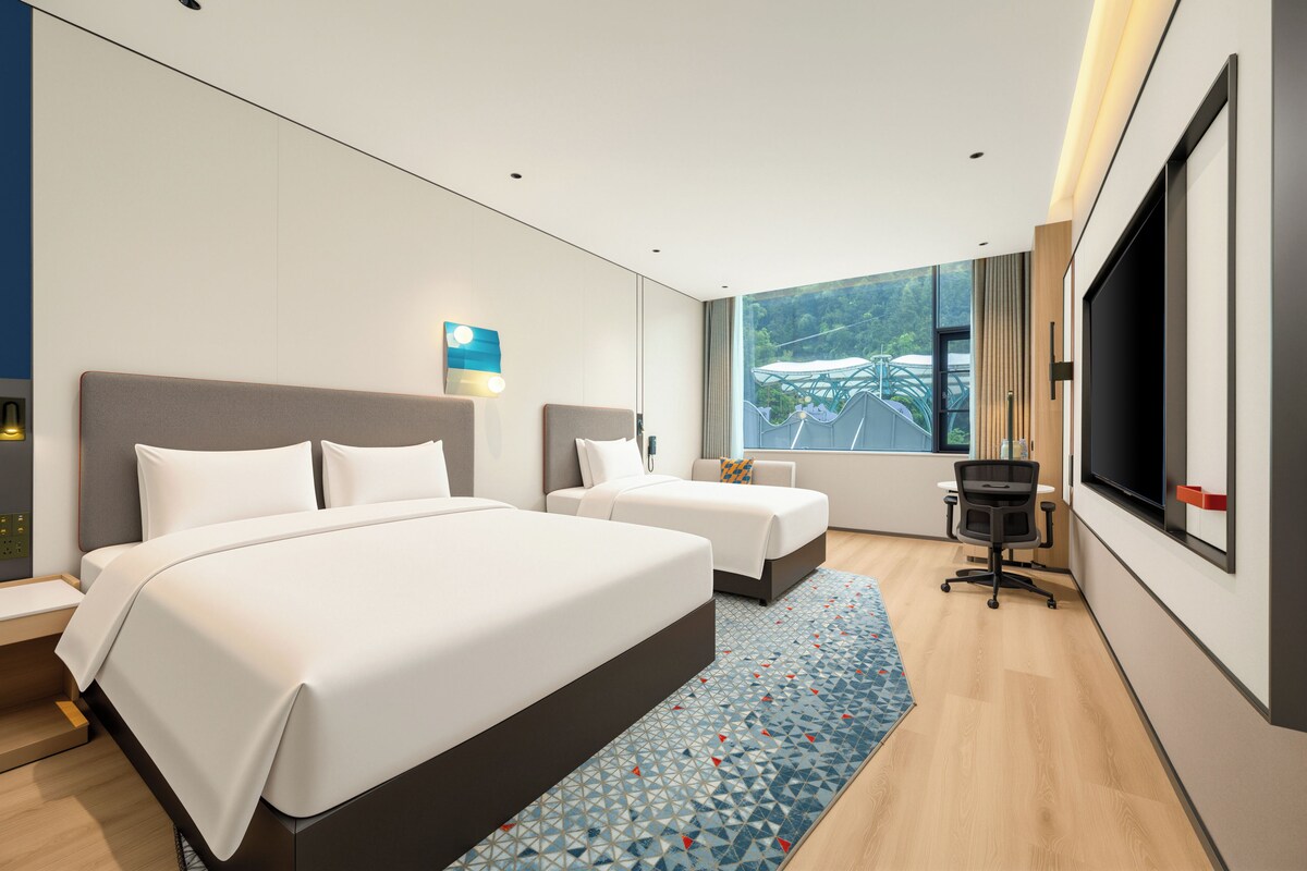 A modern hotel room with two beds, a desk, a TV, and a large window with a view of greenery.