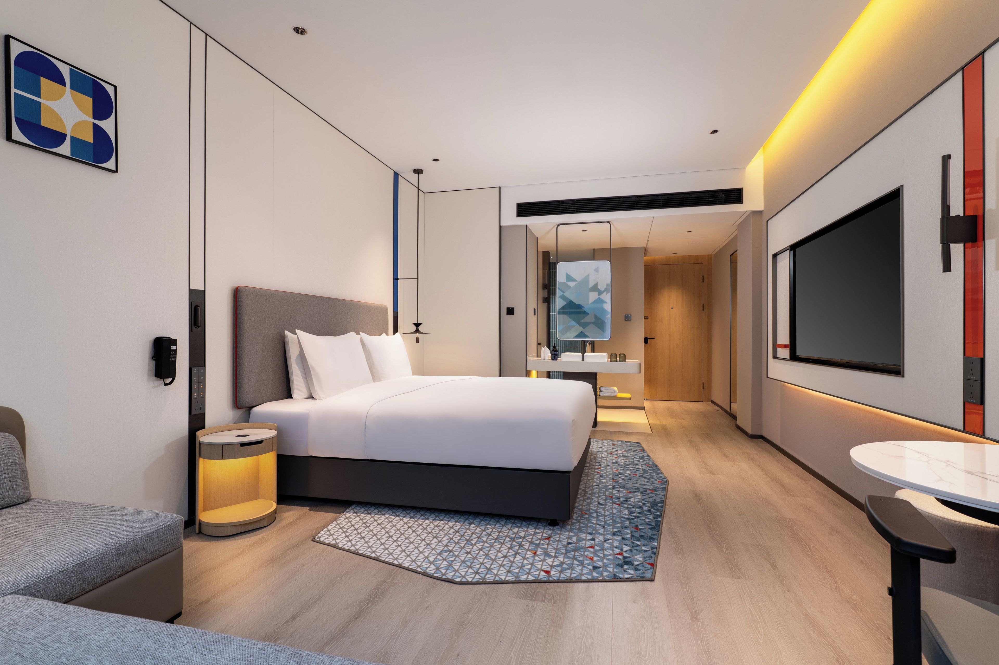 A modern hotel room with a large bed, bedside table, lamp, and a TV on the wall.