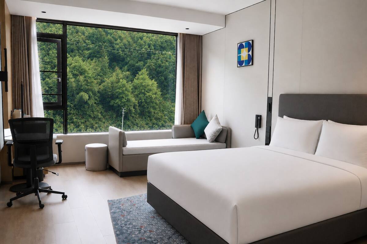 A modern hotel room with a large bed, a sofa, a desk, and a view of greenery through the window.