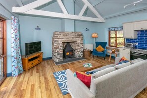 A cozy living room with a stone fireplace, wooden flooring, a blue sofa, and a kitchen with blue tiles. - Cottage with HotTub & Indoor Pool on Cornwall Farm (Searles) (Lostwithiel)