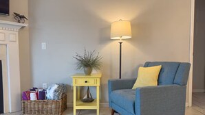 Ein Wohnzimmer mit blauem Sessel, einem gelben Beistelltisch mit Pflanze, einer Stehlampe und einem Korbkissen.