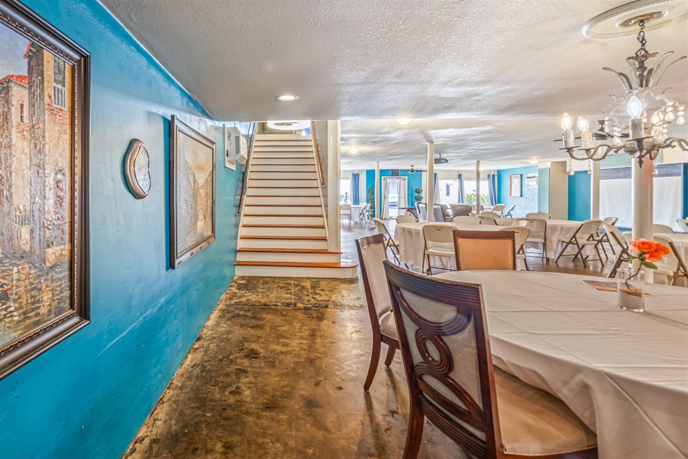 A dining area with a long table, chairs, and a chandelier. There is a staircase leading to another level.