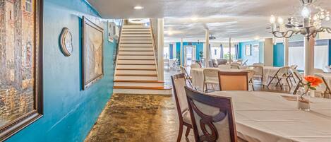 A dining area with a long table, chairs, and a chandelier. There is a staircase leading to another level.