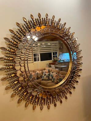 A round mirror with a gold frame featuring leaf and petal designs, reflecting a room with a sofa and a television. - Beautiful 2 bedroom apartment for rent. Close to shopping centers (Quito)