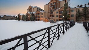 A snow-covered outdoor area with a wooden fence, buildings with multiple floors and balconies, and snow-covered trees. - Ski-in/out Marriott Mountain Valley Breckenridge sleeps 8 heated pool/hot tubs+ (Breckenridge)