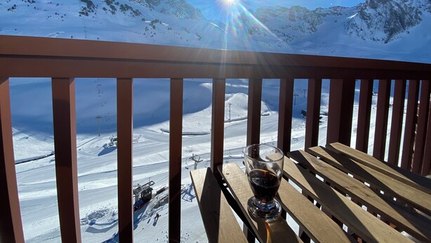 A wooden deck with a glass of drink and a bench, overlooking a snowy mountain landscape with ski slopes and a clear blue sky. - Studio at the foot of the slopes - La Mongie (Bagnères-de-Bigorre)