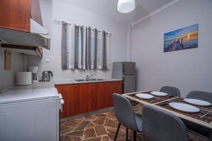 A compact kitchen with a dining area, featuring a stone-tiled floor, wooden cabinets, and a painting on the wall. - SEA GEM APARTMETS steps from the sand (Chania)