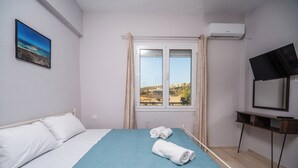 A bedroom with a bed, a window with curtains, a TV on a stand, and a painting on the wall. - SEA GEM APARTMETS steps from the sand (Chania)