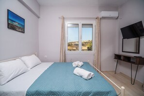 A bedroom with a bed, a window with curtains, a TV on a stand, and a painting on the wall. - SEA GEM APARTMETS steps from the sand (Chania)