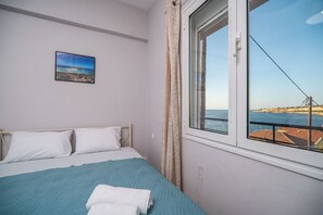 A bedroom with a bed, a window with a view of the sea, and a painting on the wall. - SEA GEM APARTMETS steps from the sand (Chania)