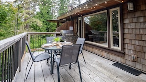 Une terrasse en bois avec une table dressée pour quatre personnes, entourée de chaises, et un barbecue situé à l’extérieur d’une grande fenêtre.