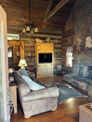A log cabin interior with a wooden ceiling, a stone fireplace, a leather sofa, and a television. - Rustic Farm Retreat: 2BR Cabin Amidst Rolling Fields & Fresh Country Air. (Spartanburg)