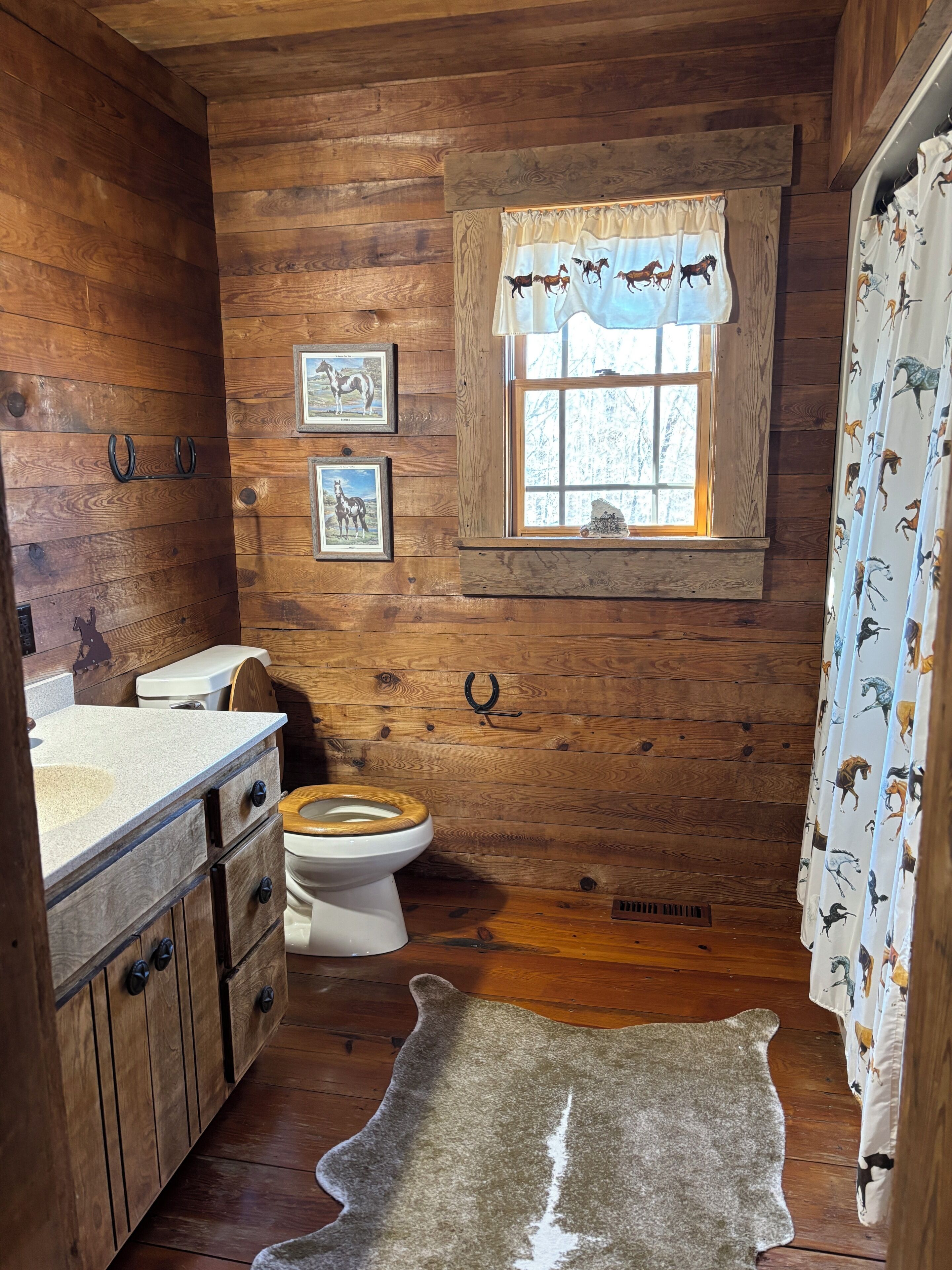 A rustic cabin bathroom with wooden walls, a wooden toilet, a window with a horse-themed curtain, and a rug on the floor.