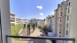 Blick von einem Balkon auf eine enge Straße mit mehrstöckigen Gebäuden, einem klaren Himmel und einem sichtbaren Logo in der Ecke.