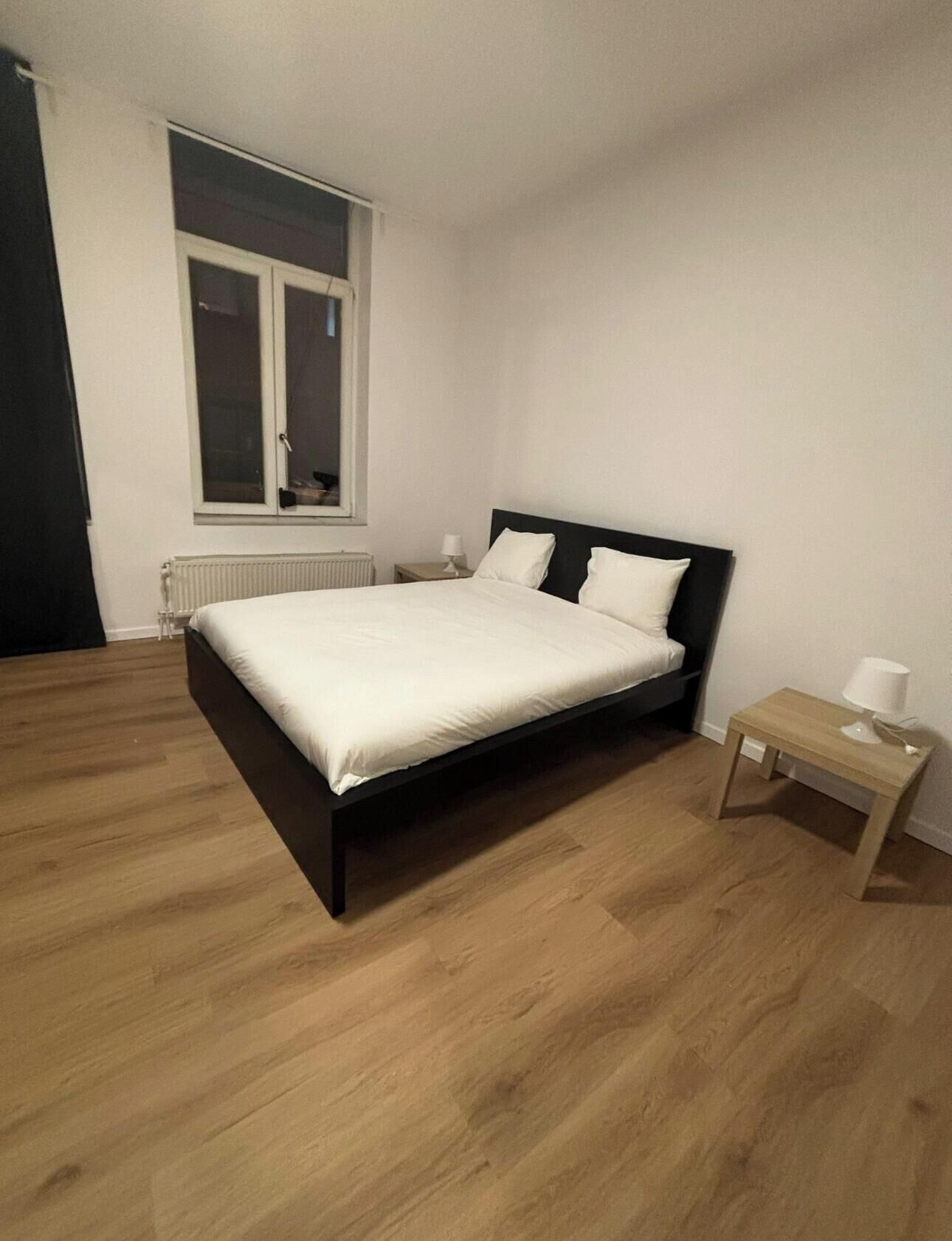 Un dormitorio con cama, mesita de noche, ventana y radiador.
