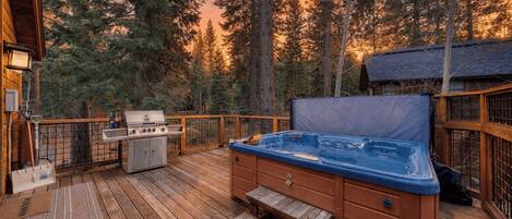 A wooden deck with a hot tub, grill, and outdoor seating area surrounded by trees.