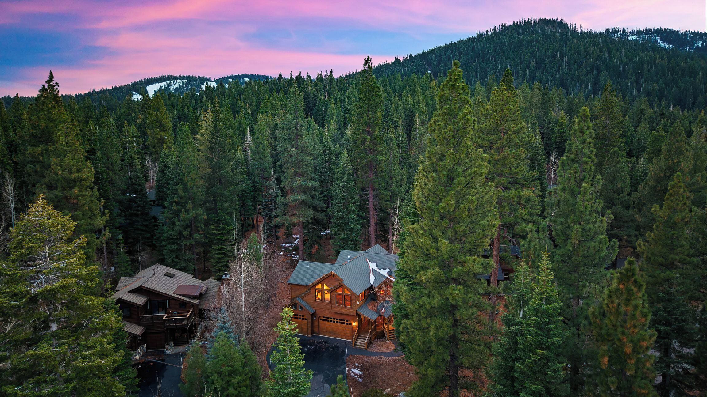 A cabin with a large deck and a garage, surrounded by dense forest and mountains.