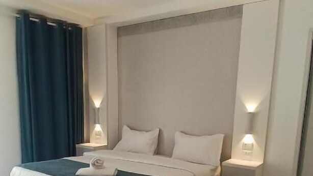 Una cama bien hecha con ropa de cama blanca y aguamarina, dos mesitas de noche con lámparas y una silla con estructura metálica.