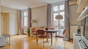 Ein geräumiges Wohnzimmer mit Essbereich, großen Fenstern mit Vorhängen, einem Flachbildfernseher und einer Heizung.