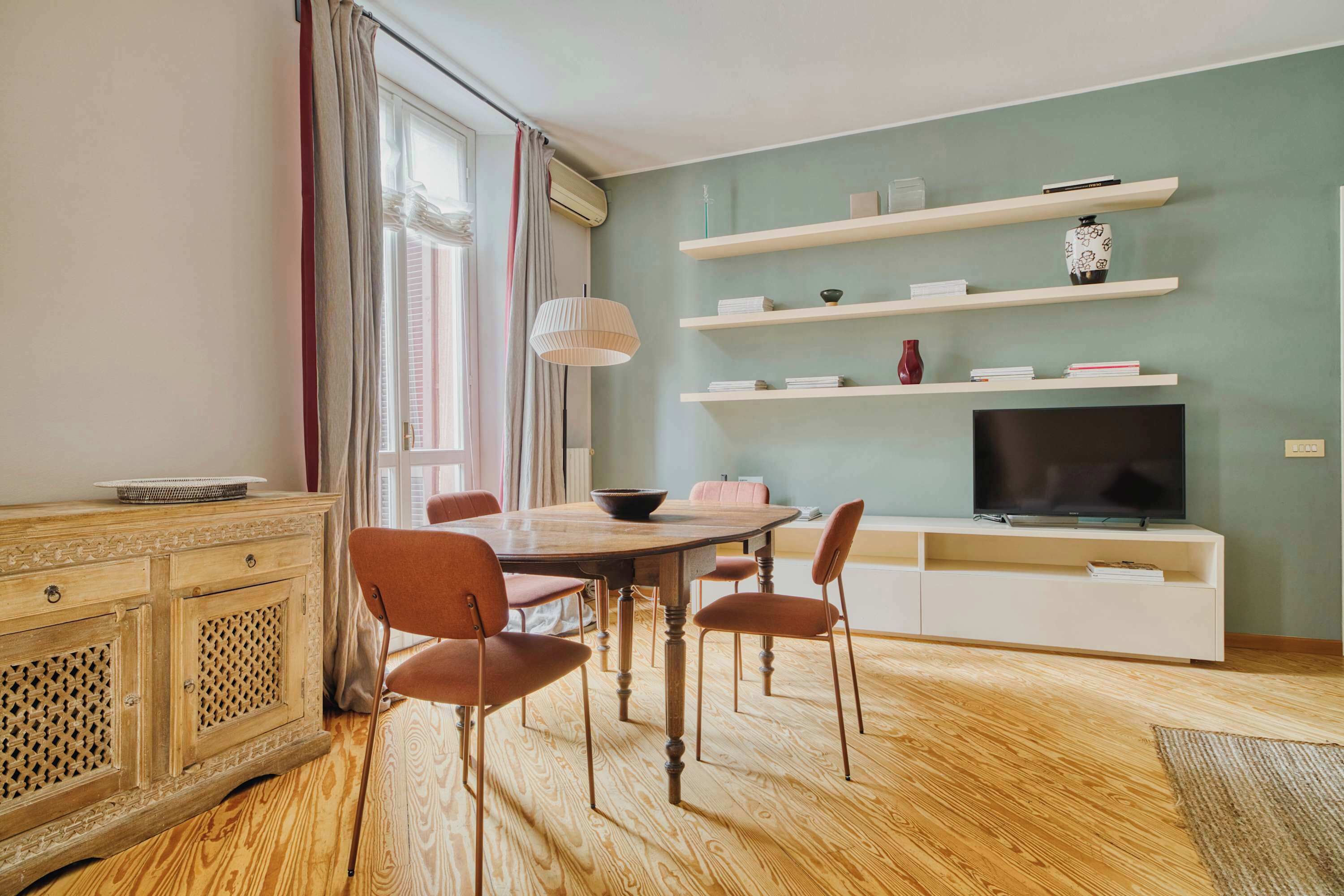 Ein modernes Wohnzimmer mit einem Holzespeisetisch, vier Stühlen, einem Fernseher auf einem weißen Schrank und Regalen mit Dekoration.