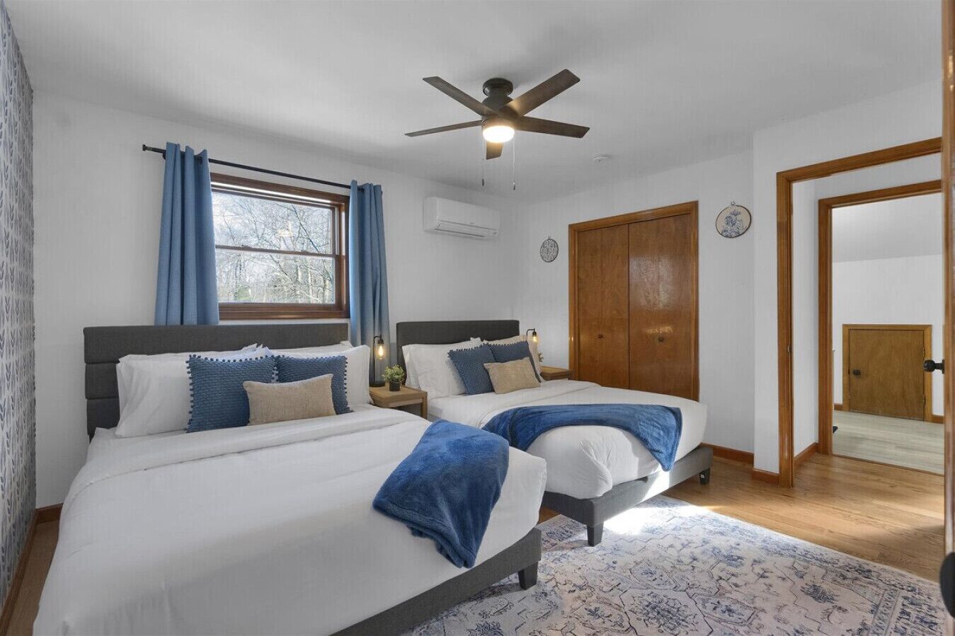 Une chambre avec deux lits, un ventilateur de plafond, une fenêtre avec des rideaux, une armoire en bois et un tapis à motifs.