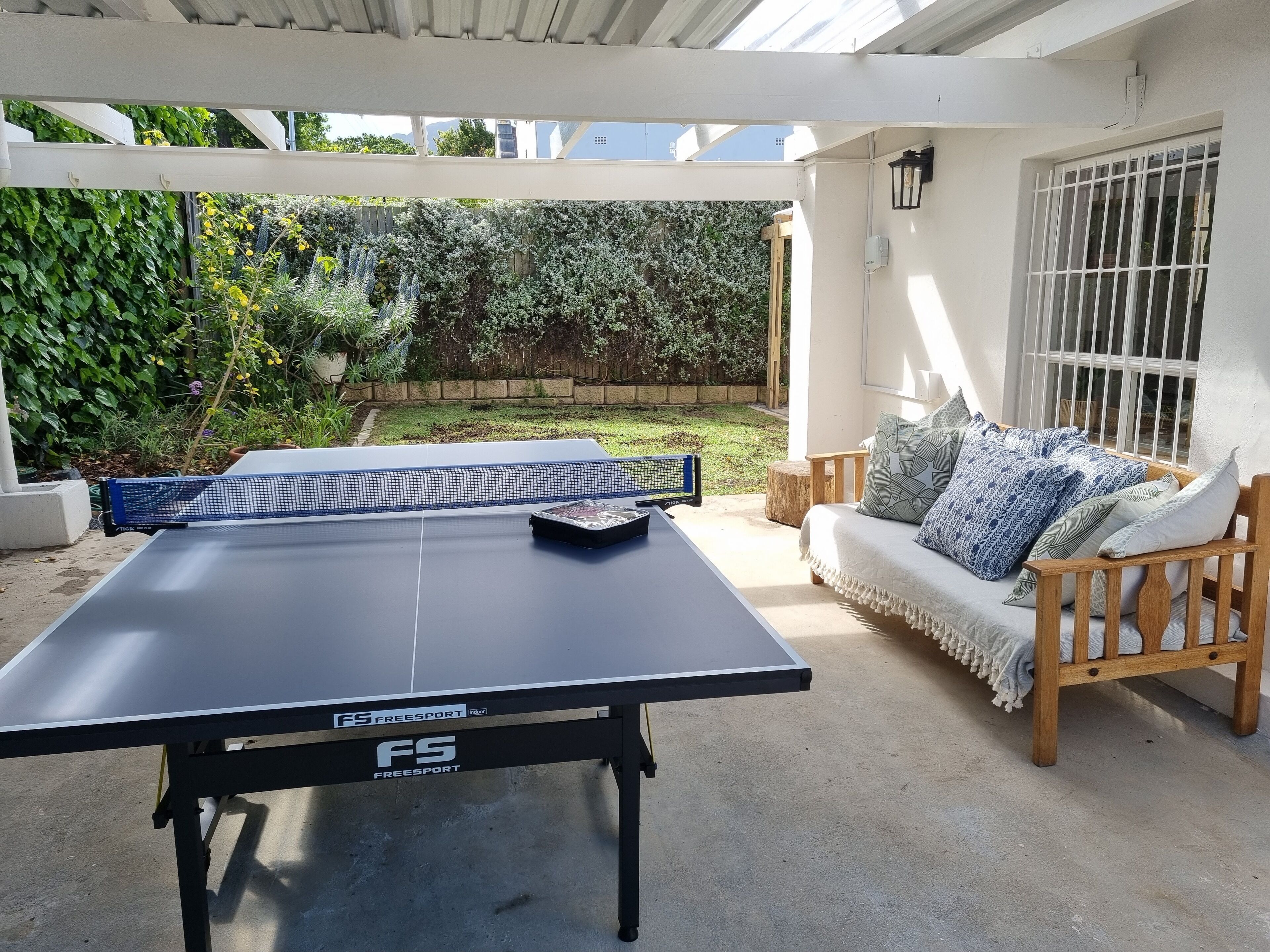 An indoor ping pong table with a net, a wooden bench with cushions, and a view of a garden with a white wall and a window with bars.