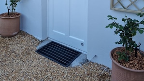 Puerta blanca con un panel de vidrio, flanqueada por dos faroles de pared y una ventana celosíaca a la izquierda. Hay dos plantas en macetas, una a cada lado de la puerta.