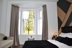 Ein Schlafzimmer mit einem Bett, einem Sessel, einem Nachttisch mit Lampe und einem Fenster mit Vorhängen.