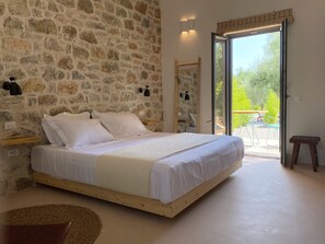 A bedroom with a stone wall, a wooden bed, a small stool, and a view of the outdoors through a glass door. - Inspiro Retreat (East Mani)