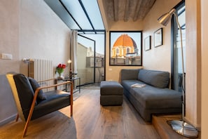 A modern living room with a sofa, ottoman, and a chair, a radiator, a table with a vase, and a framed picture of a building. - San Lorenzo Terrace Apartment (Florence)