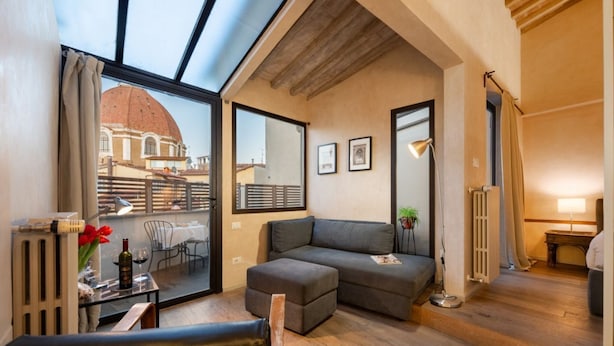 A modern living room with a sofa, a table with a bottle and glasses, and a view of a building through large windows. - San Lorenzo Terrace Apartment (Florence)