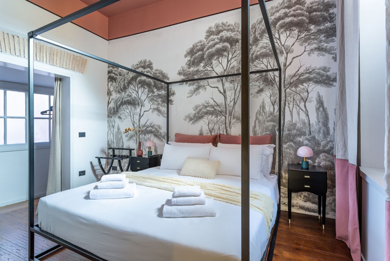A bedroom with a four-poster bed, a nightstand, a lamp, and a mural of trees on the wall.