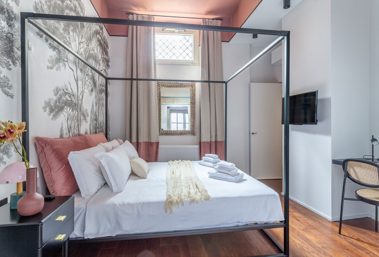 A four-poster bed with white linens, a beige throw blanket, and a set of towels. A television is mounted on the wall to the right.
