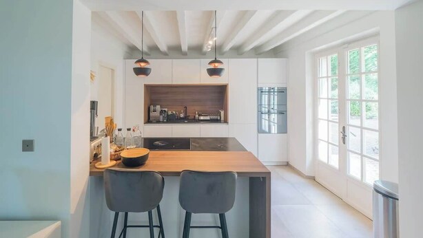 Une cuisine moderne dotée d’un îlot central, de deux tabourets de bar, d’un éclairage suspendu et offrant une vue sur l’extérieur grâce à de grandes fenêtres.