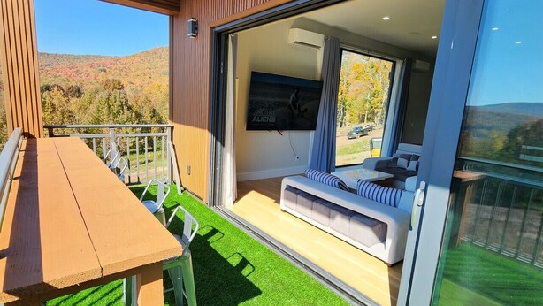 A modern outdoor living area with a wooden table and chairs, a sofa, and a large sliding glass door leading to a bedroom with a bed and a mounted TV. - Modern Catskills Mountain Retreat with Rooftop Hot Tub on 22 Private Acres (Denver)