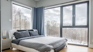A modern bedroom with a large bed, a nightstand, and a window with a view of trees. - Modern Catskills Mountain Retreat with Rooftop Hot Tub on 22 Private Acres (Denver)