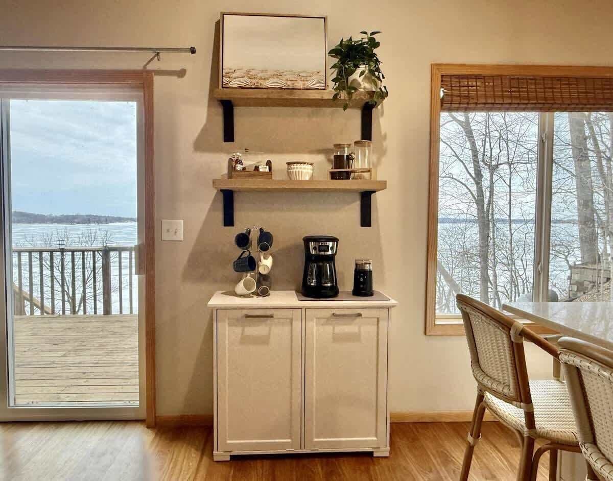 Een kamer met houten vloer, een eettafel met stoelen, een kast met keukenbenodigdheden, een raam met uitzicht op bomen, en een deur naar een balkon.
