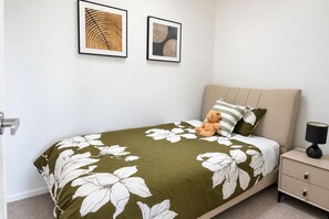 A bedroom with a bed, a nightstand, a lamp, a teddy bear, and two framed artworks on the wall.