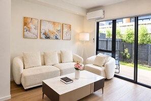 A living room with a beige sofa, a coffee table with a book, and a vase with flowers. There are three abstract paintings on the wall and a window with a view of a backyard.