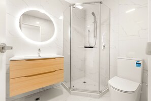 A modern bathroom with a glass-enclosed shower, a round mirror, a white toilet, and wooden cabinetry. - Family-Friendly New 3BR Townhouse | Gated Community & Secure Parking (Glen Eden)