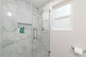 A glass-enclosed shower with a handheld showerhead, a small shelf with toiletries, and a window with frosted glass. - Charming 4-bedroom+4 bath (Houston)