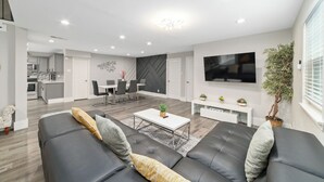 Ein modernes Wohnzimmer mit Sofa, Couchtisch und Fernseher. Es gibt einen Essbereich mit Stühlen und einem Tisch.