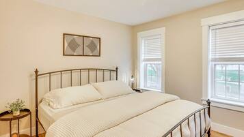 A bedroom with a metal bed frame, a large window with blinds, a bedside table with a lamp, and a wall-mounted artwork.