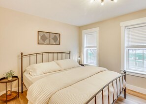 Un dormitorio con estructura de cama metálica, un ventanal con persianas, una mesita de noche con lámpara y una obra de arte colgada en la pared.