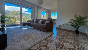 Ein geräumiges Wohnzimmer mit einer großen Eckcouch, einem Couchtisch und Blick auf eine gebirgige Landschaft durch große Fenster.