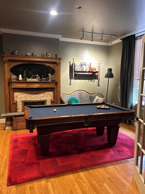 A pool table with a red rug, a fireplace, and a bookshelf with books and decorative items.